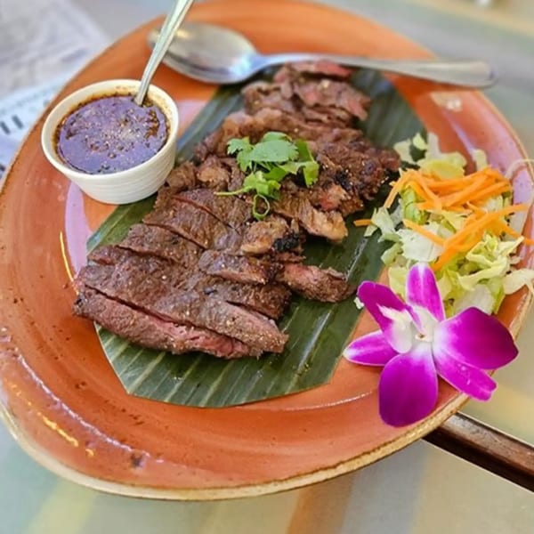 Sliced grilled steak on a banana leaf with dipping sauce, salad, and an orchid.